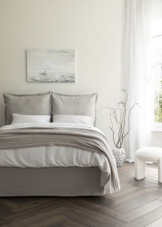 A white bedroom with herringbone flooring, a soft grey bed, and a white boucle stool