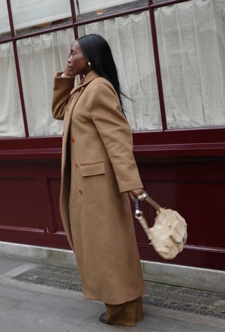 A woman wearing a long camel coat with brown trousers and an off-white handbag.