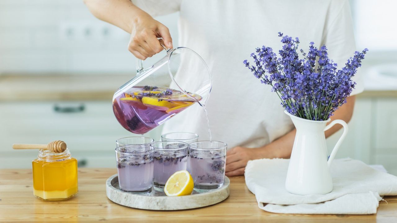 A woman pours lavender tea into five small cups next to a jar of honey and a pitcher of lavender flowers