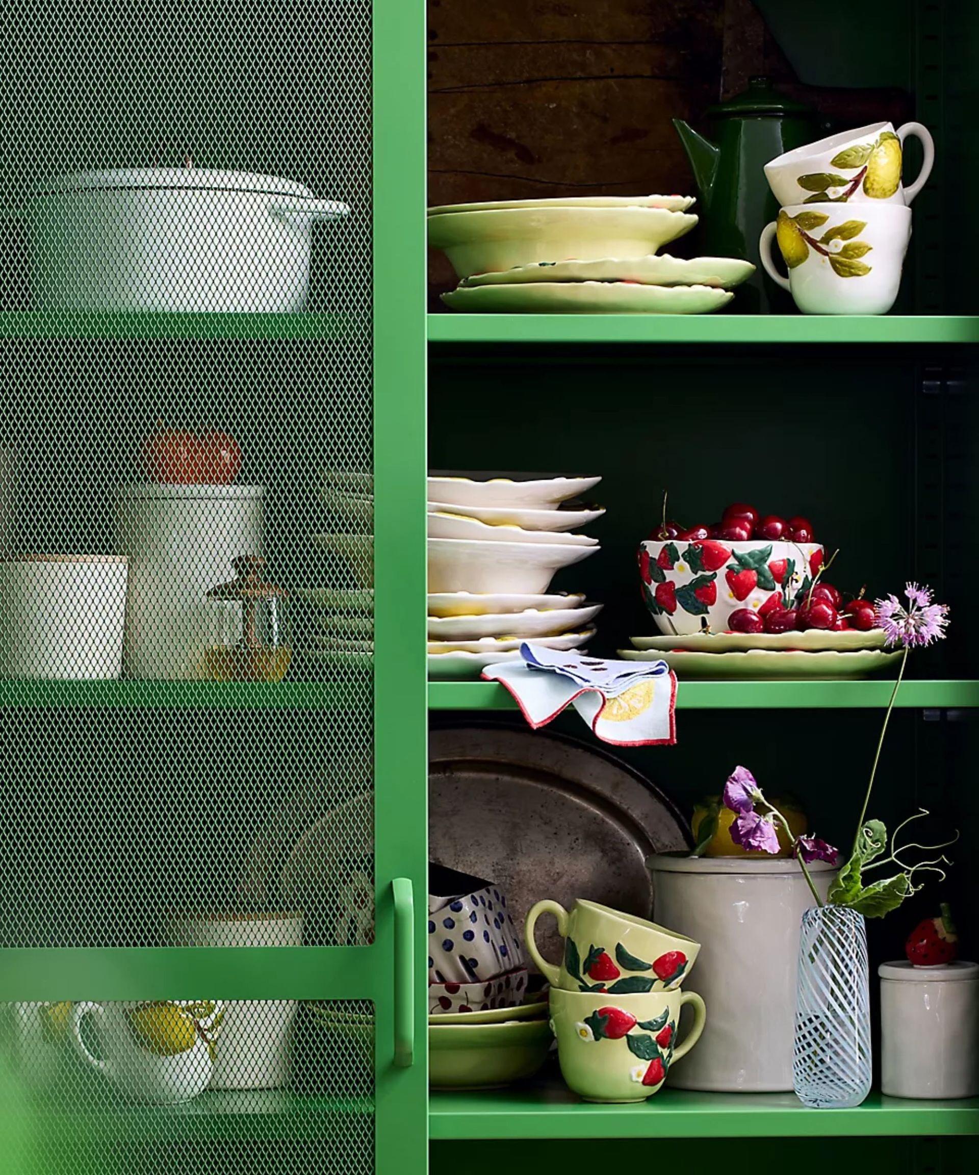 green kitchen cabinet with shelves filled with anthropologie fruit themed kitchenware, crockery, and decor