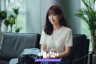 a woman (kang han-na) smiles while sitting on an office couch, in the kdrama 'frankly speaking'