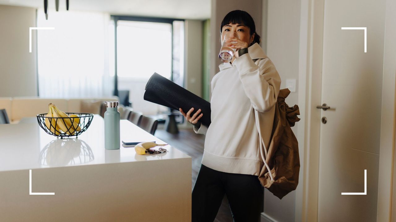 Woman coming into a kitchen, holding a yoga mat and glass of water, about to put workout clothes in washing machine