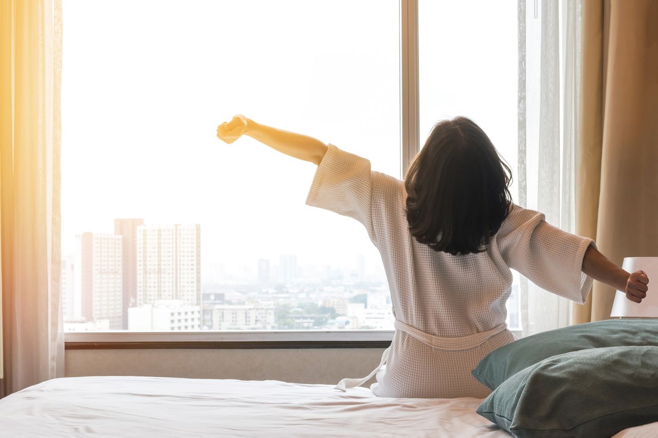 A woman in a robe stretches her arms from a bed in front of a large window