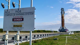 An orange rocket with a white top stands against a dynamic sky. A danger sign stands on the left.