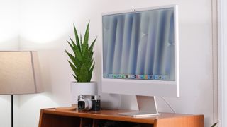 Apple iMac on a wooden desk against a white wall next to a lamp, camera and green plant