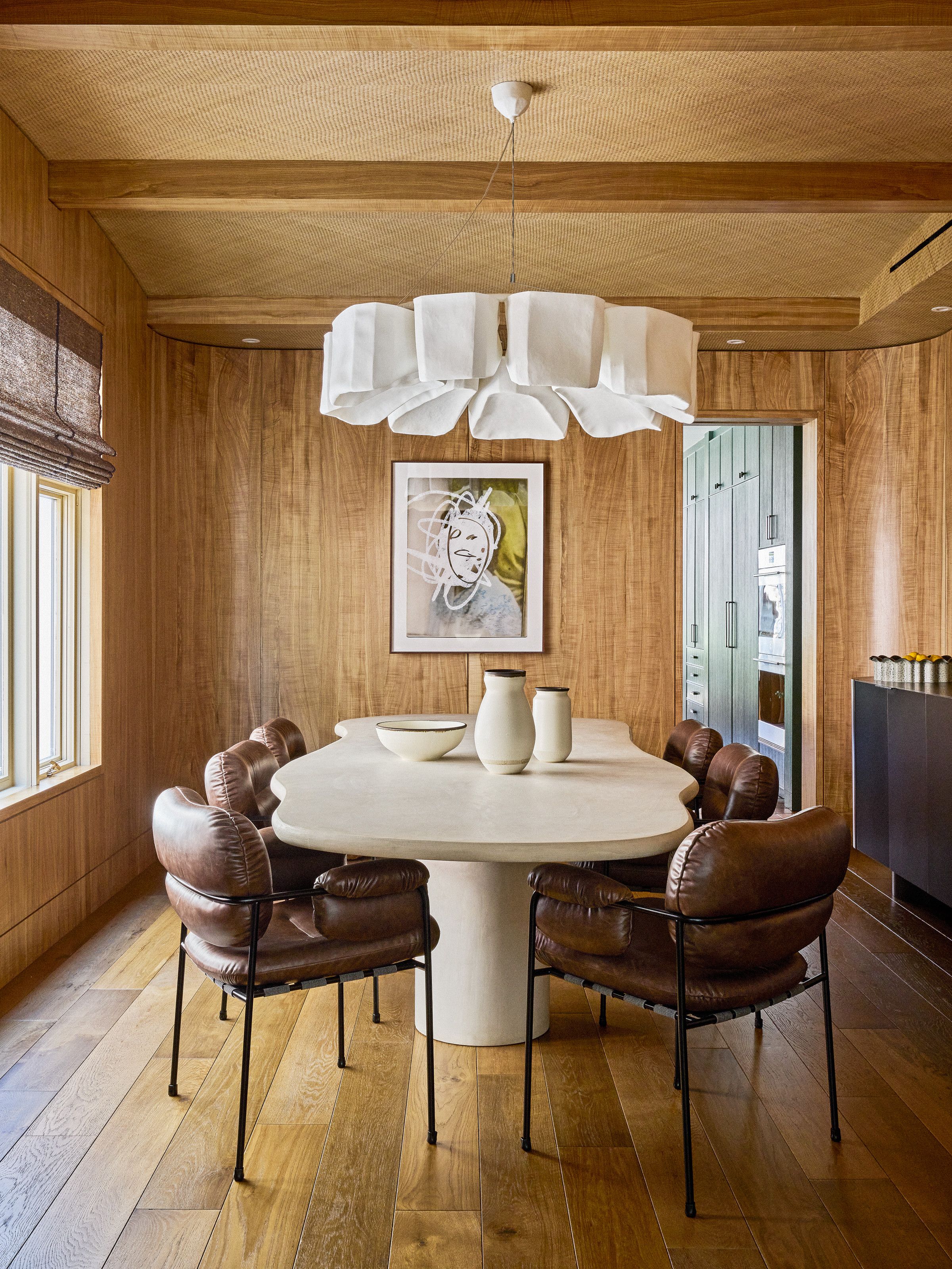 Dining room with wood panelled walls, ceiling with beams, wood floor, curved travertine table, brown leather chairs and oversized fabric chandelier