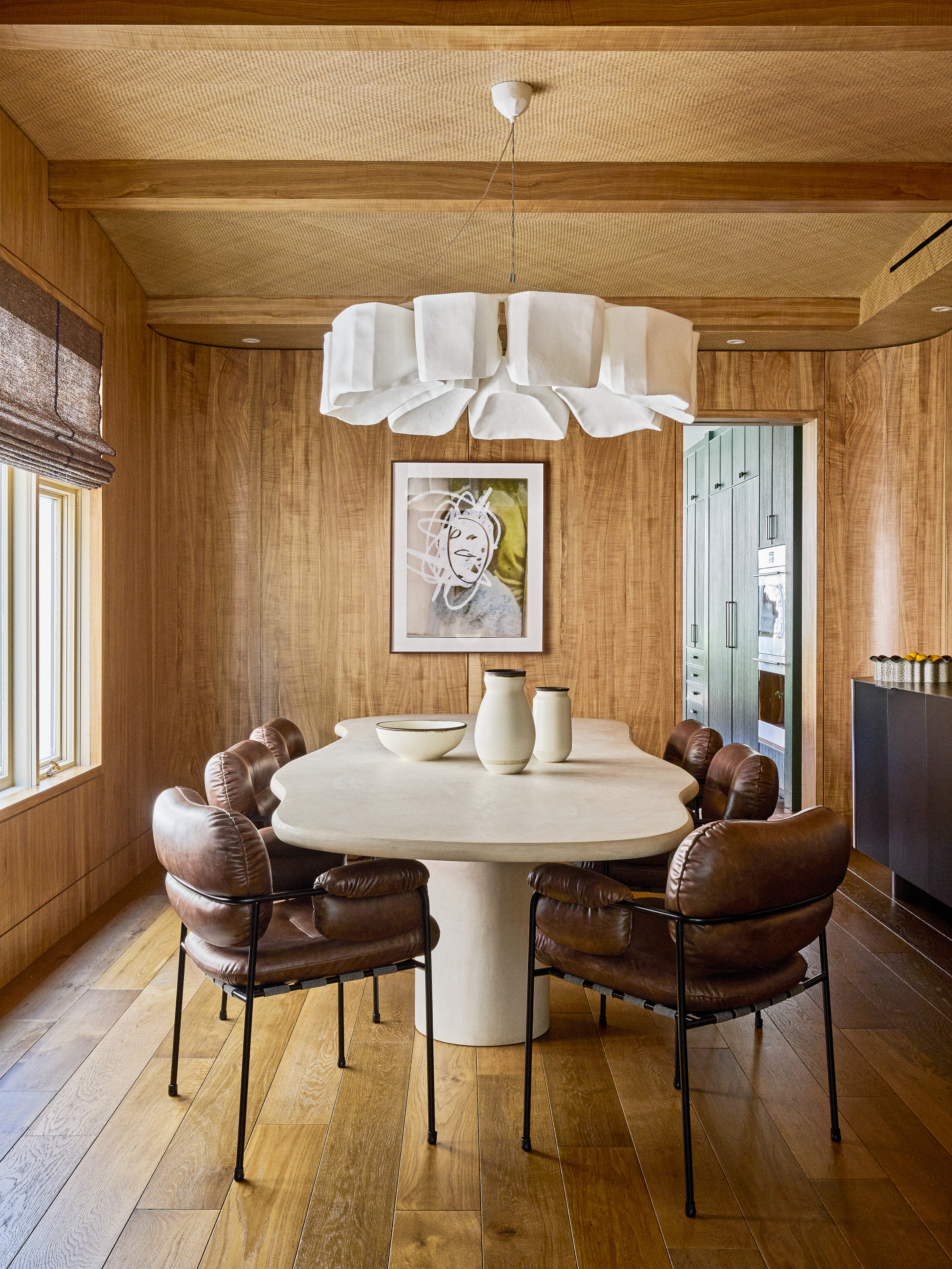 Dining room with wood panelled walls, ceiling with beams, wood floor, curved travertine table, brown leather chairs and oversized fabric chandelier