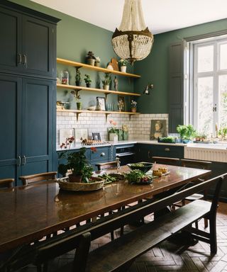 Navy kitchen diner with green walls, open shelving, long table and benches, metro tile backsplash, ribbed sink, open shelving, chandelier,