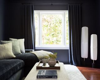 Black painted living room with black curtains and sofa, dark wood flooring with lantern style white floor lamps, cream rug, cushions and footstool