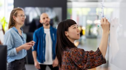 Agile development concept image showing female developer writing down strategy points on a whiteboard with male and female colleagues pictured in background.