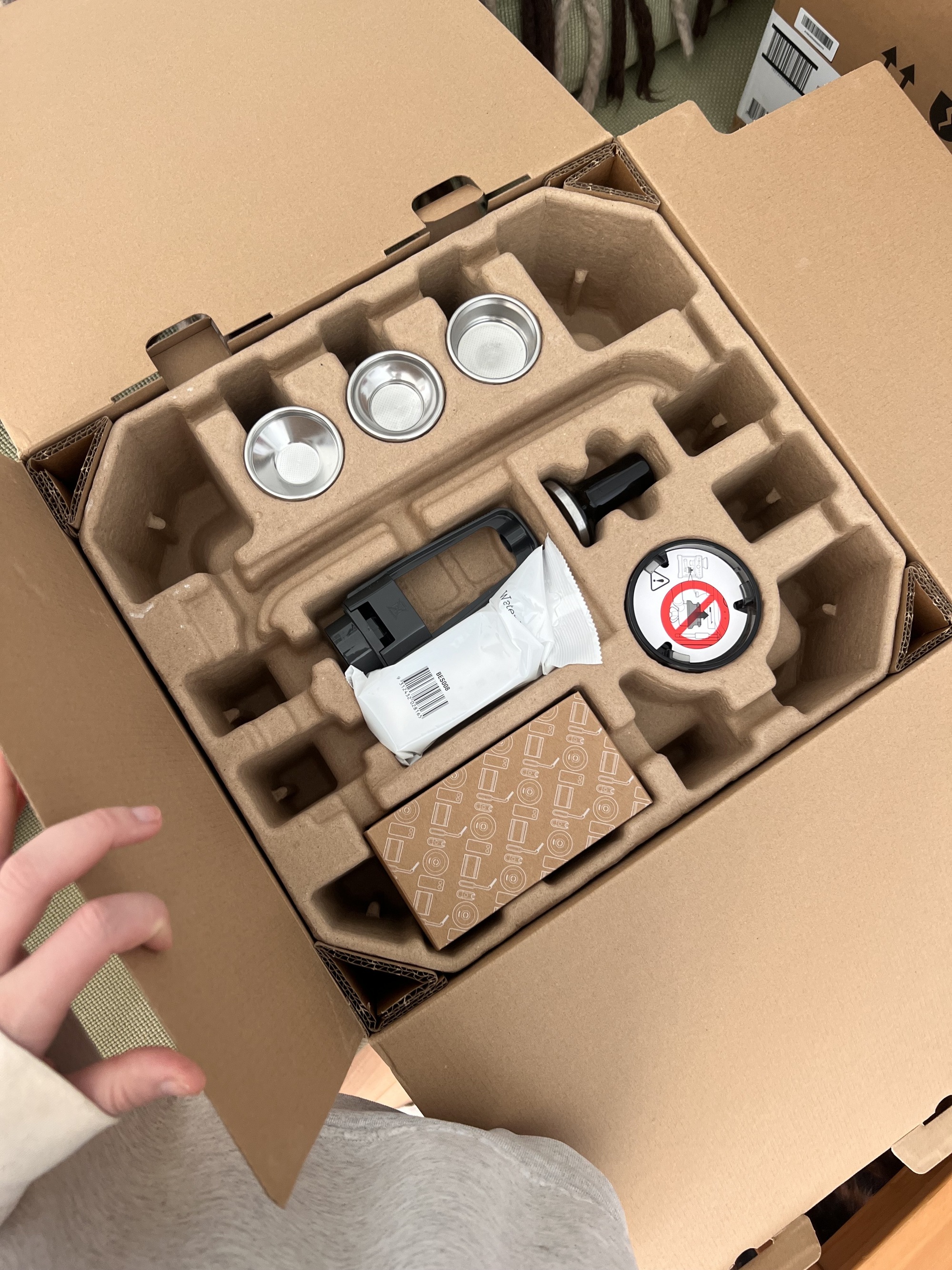 A hand opening a cardboard box that has a coffee machine and other coffee machine accessories inside.