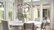 An all white and cream dining space with glass doors surrounding, and a round white dining table and miss matched white chairs, with a circular chandelier above.