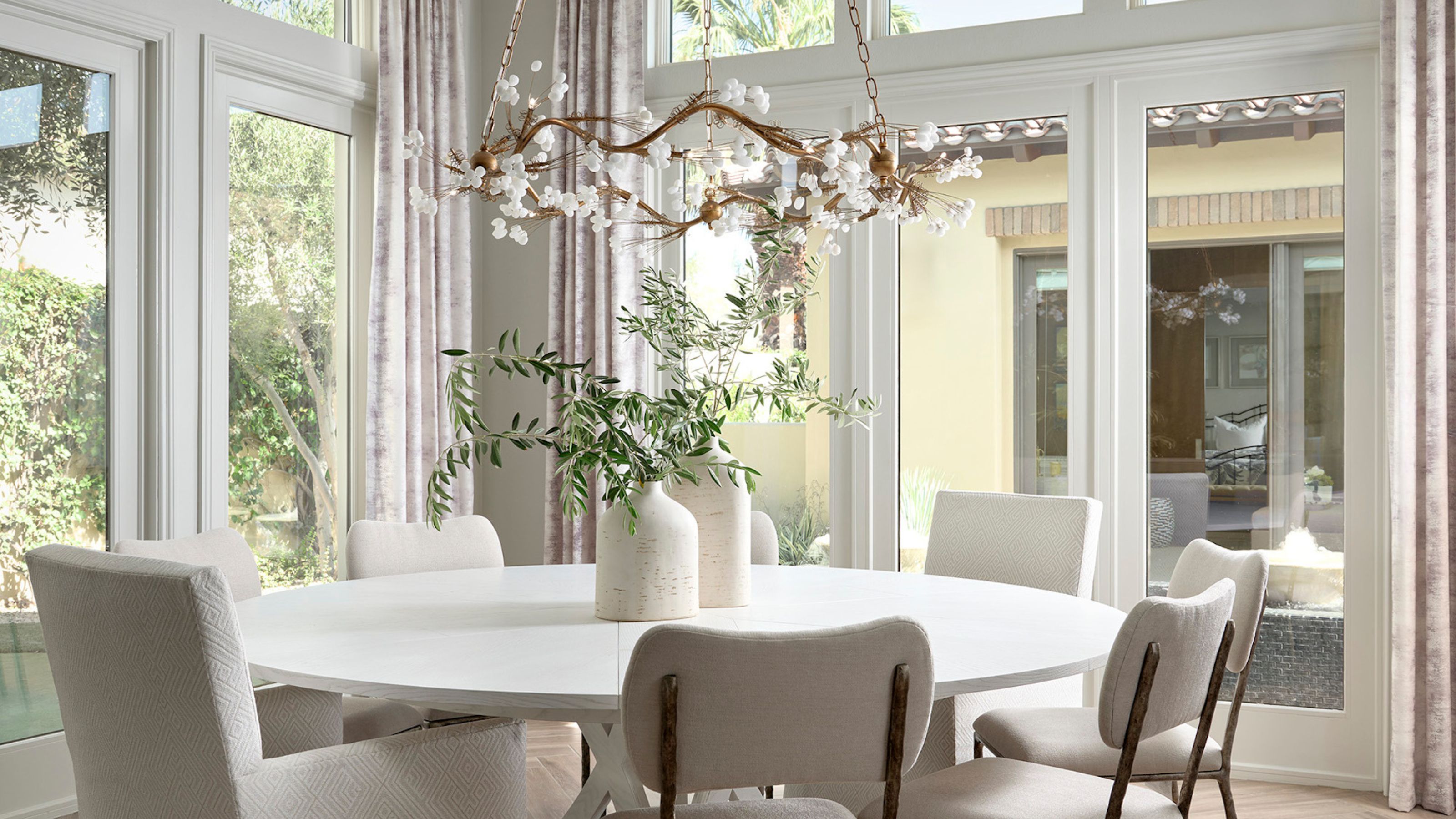 An all white and cream dining space with glass doors surrounding, and a round white dining table and miss matched white chairs, with a circular chandelier above. 