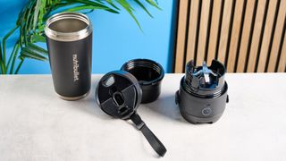 the Nutribullet Flip with a black insulated tumbler and a portable battery-powered blade photographed with a blue background. The tumbler has a carry strap and a sippy lid you can interchange, and an on/off button on the base.