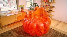 A bright orange JELL-O shaped inflatable chair in an apartment with pink walls, wooden furniture, and a gray rug