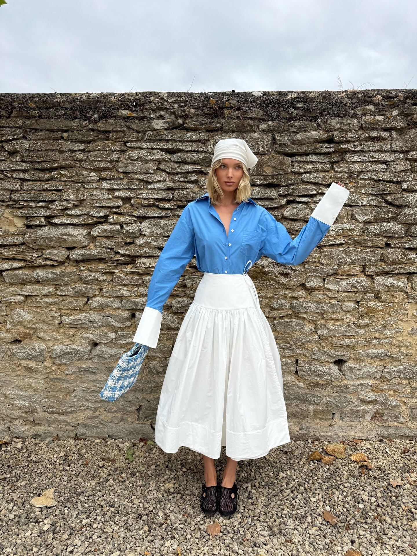 Elsa Hosk wearing a head scarf, Helsa blue button-down shirt and white poplin midi skirt, and Alaia fishnet slippers in the Cotswolds