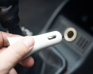 A person holding a white plug with a car's 12v plug socket in background on a soft focus