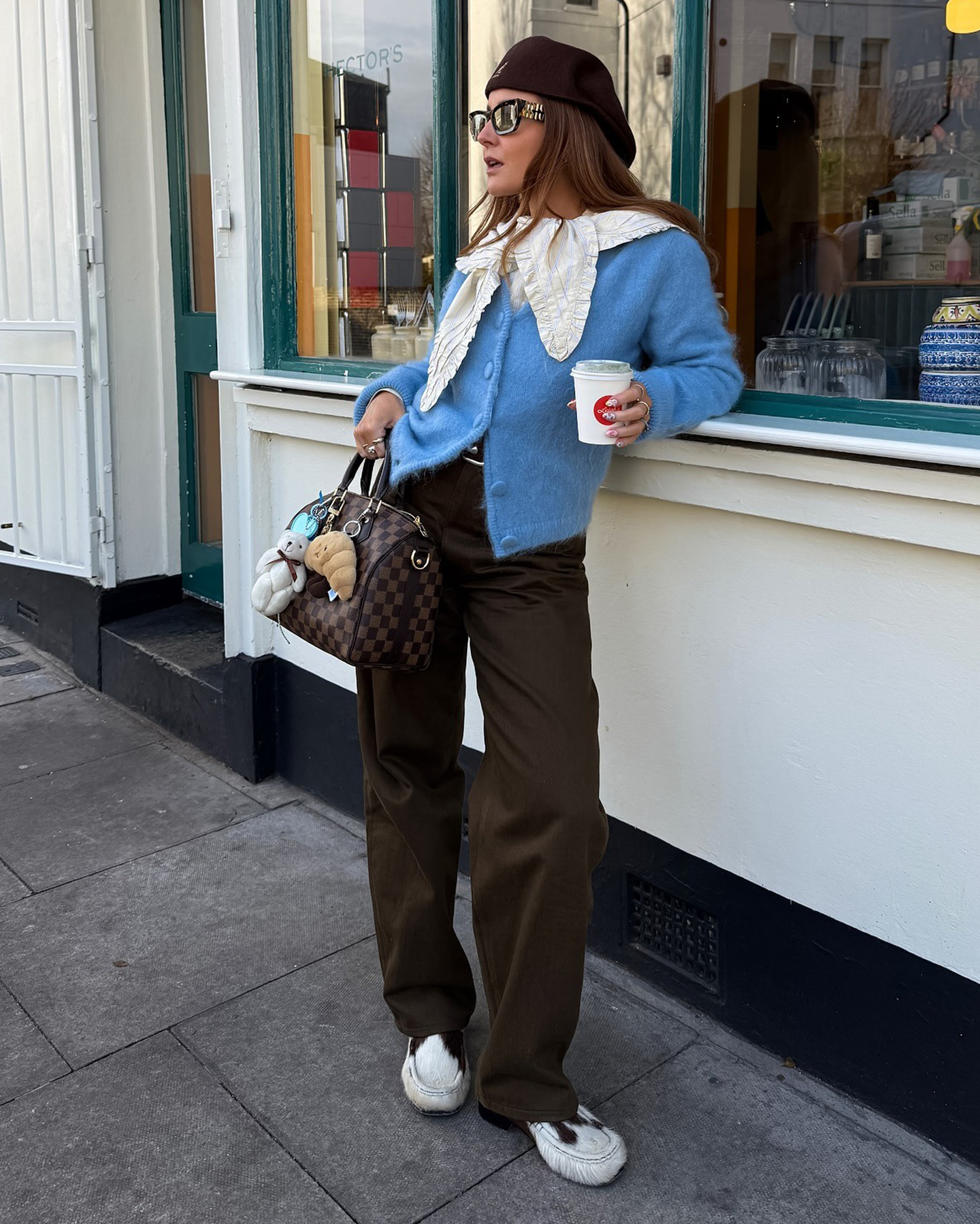 British style influencer Rebecca Ferraz Wyatt poses on a London sidewalk holding a coffee wearing a brown Kangol hat, ruffle collar, light blue cardigan sweater, a Louis Vuitton bag with bag charms, brown jeans, and cow-print loafers.