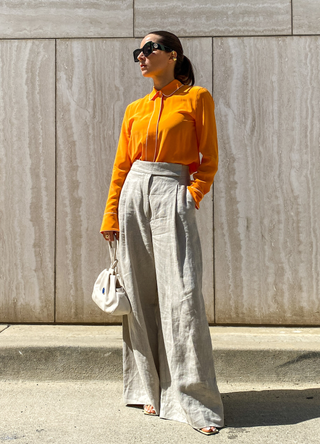 Julia Comil wears a bright orange silk blouse with white piping detail, long sleeves by Equipment, beige linen high waisted wide trousers oversized pants by Nehera Official, cream square toe sandals by Wandler, a white eggshell wristlet bag in leather with blue agate stone by Mehry Mu, green tortoise cateye oversized sunglasses by Lanvin, during an online remote street style fashion photo session via Apple iphone / Facetime and the CLOS app as the model is located in Beverly Hills - California and the photographer in Paris - France, on June 04, 2021 in Paris, France