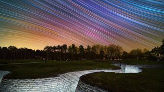 A white-bricked trench lays recessed in the pale green grass of a World War I memorial in France. Above, the night sky is diagonally streaked with a rainbow of stars.