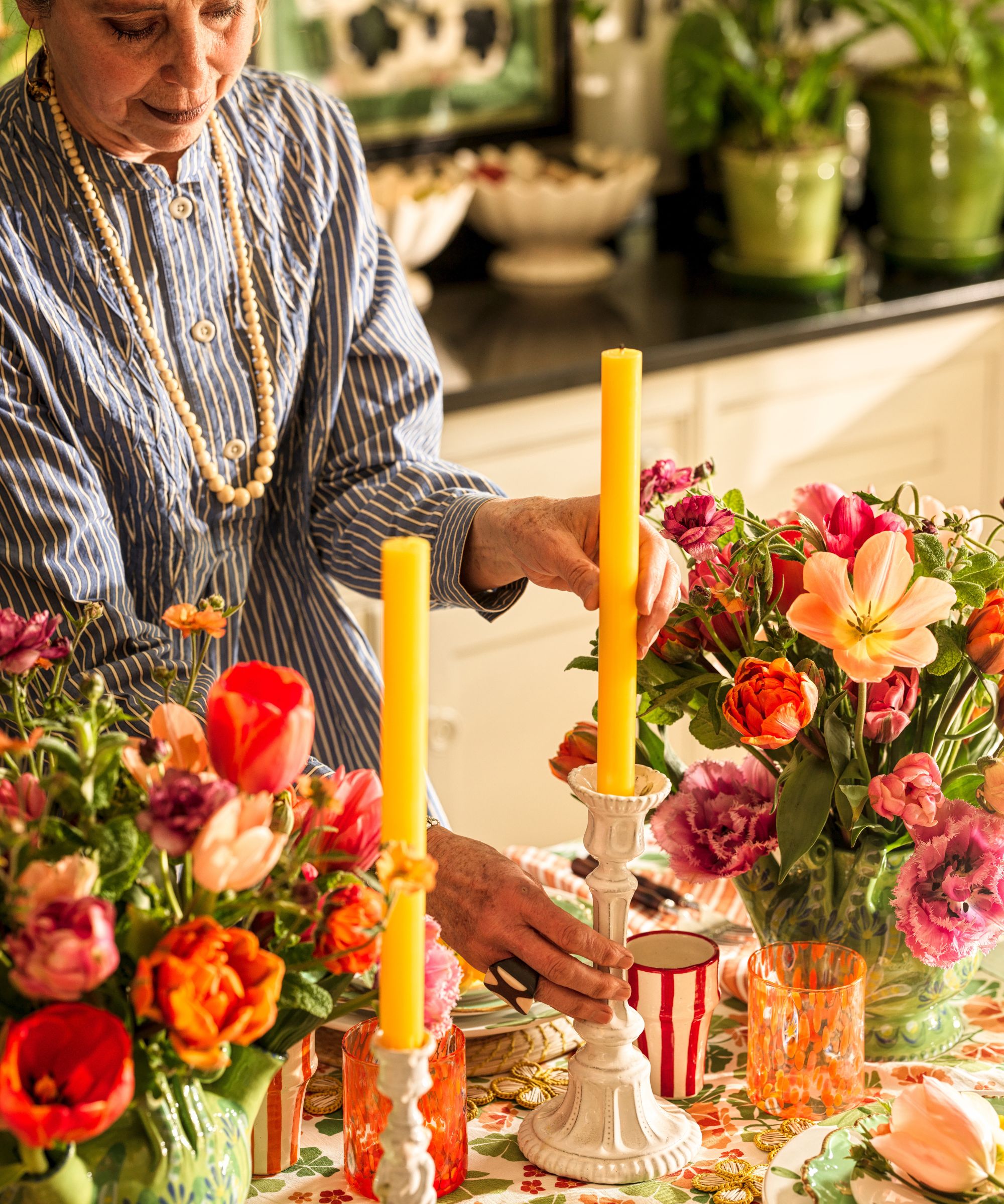 Butter Wakefield, wearing a blue striped shirt and pearl necklace, carefully adjusts a tall yellow taper candle in a white ceramic holder on the decorated dining table