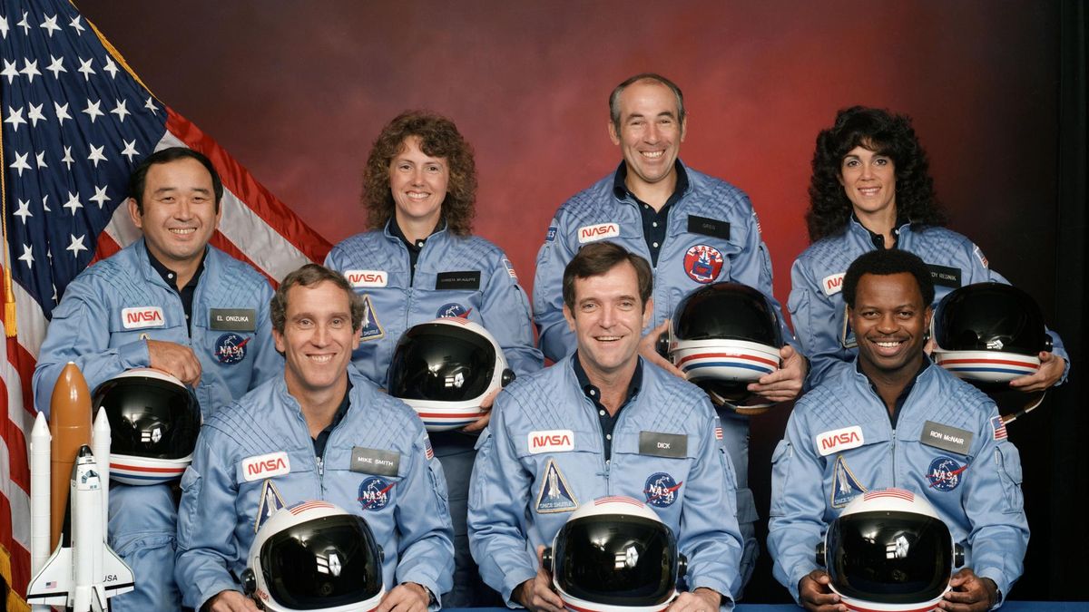 seven astronauts in blue flight suits sit for a portrait while holding their helmets