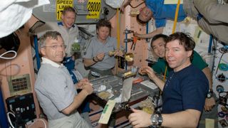six male astronauts wearing gray, blue or green polo shirts and tan slacks float around a table, enjoying a meal aboard a space station