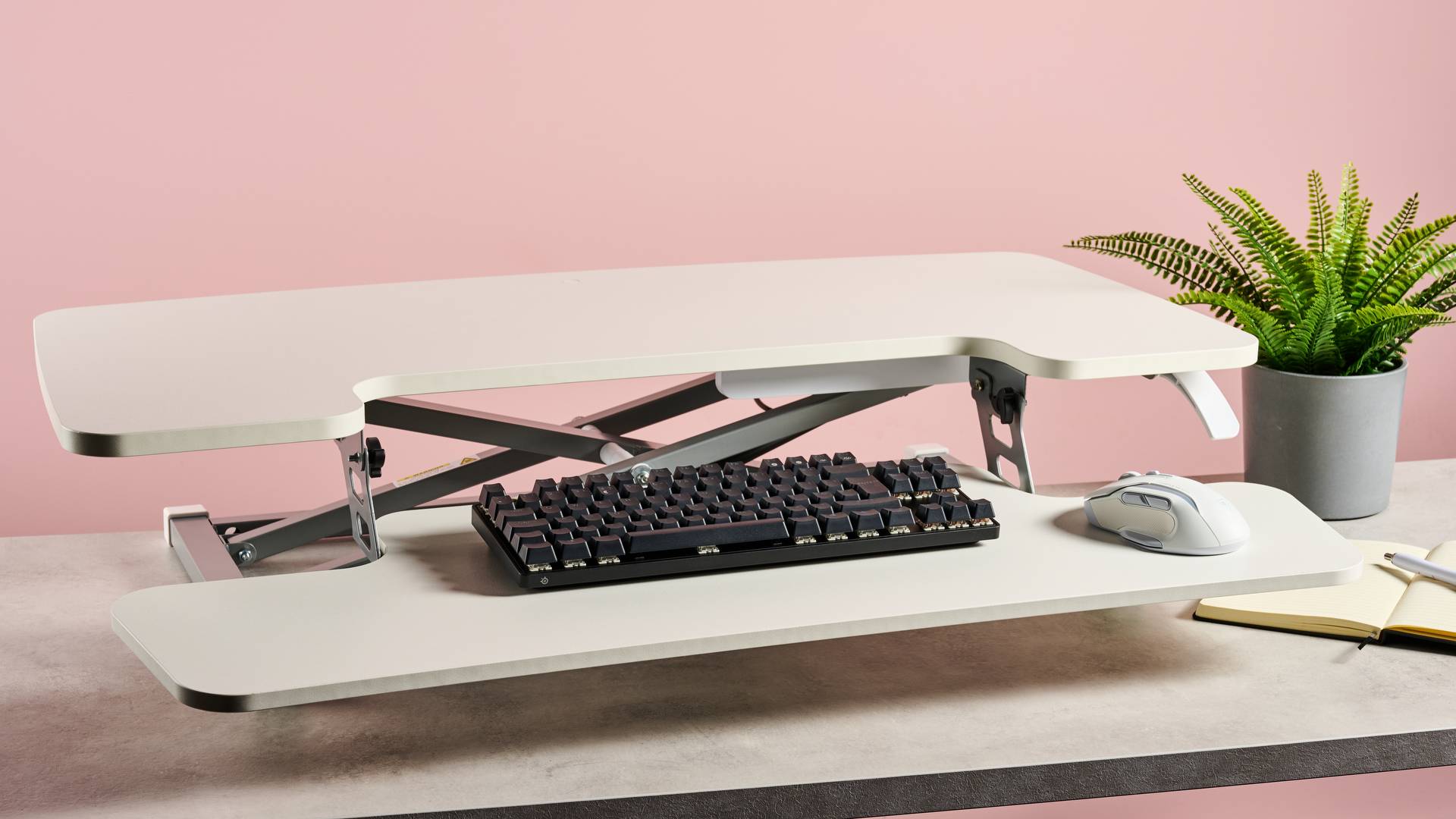 Flexispot M17 on a stone-effect desk in front of a pink background. There is a keyboard and mouse on the table, and a potted plant and notebook on the right.