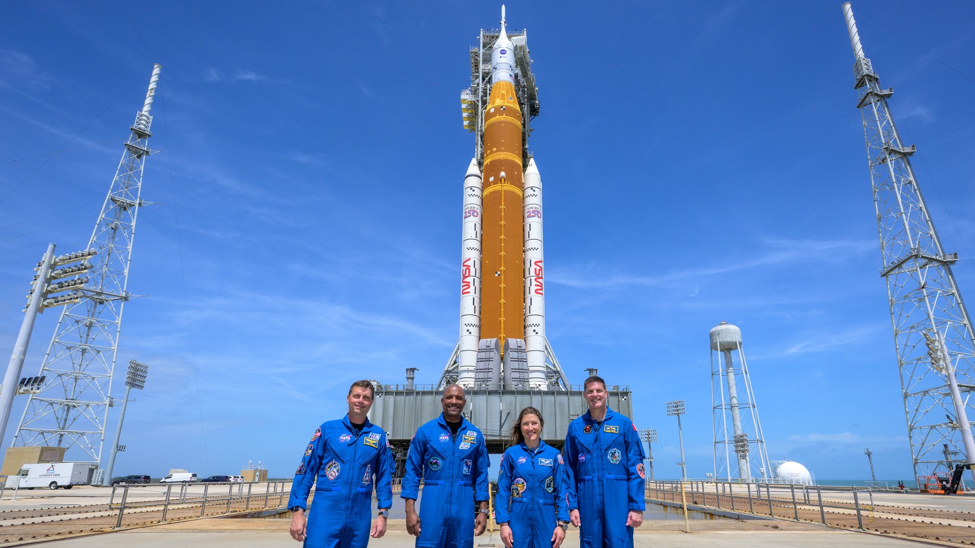 four people in blue flight suits in front of a rocket on a launch pad