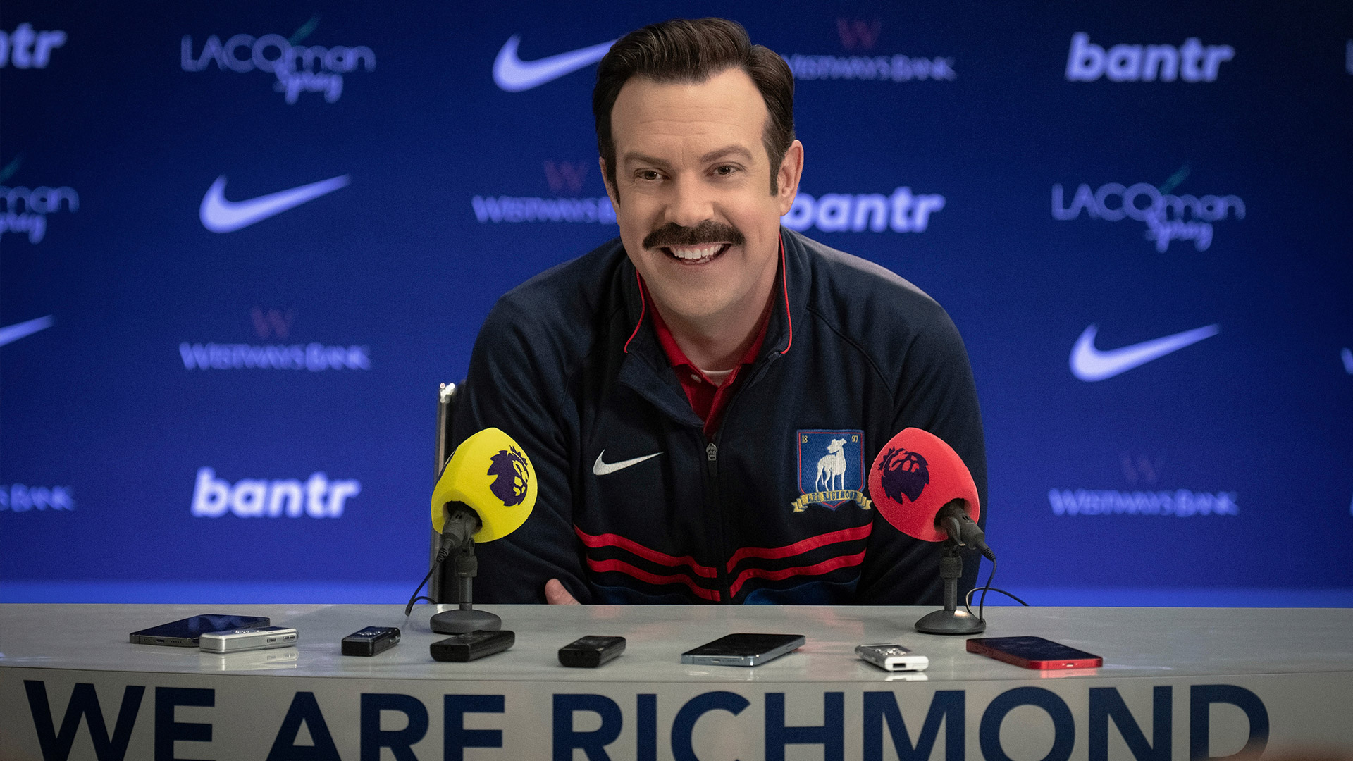 Jason Sudeikis sits at a table with microphones in front of him