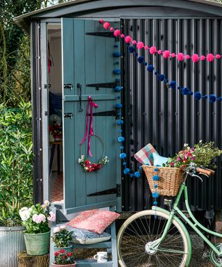 Storage ideas for sheds illustrated by a black painted corrugated iron shed with colorful pom pom garlands and a vintage bicycle.