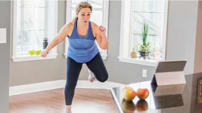 woman facing the camera wearing a blue vest and dark leggings doing an aerobics move in front of a digital device resting on a countertop with windows behind her