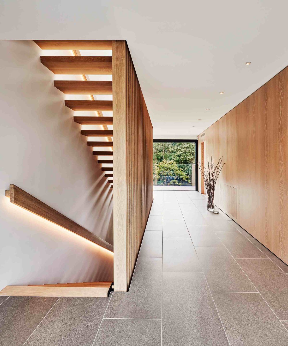 Modern hallway of a Baufritz home, featuring a timber floating staircase, a timber-clad wall, large floor tiles and a large picture window at the end