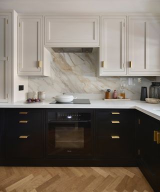 Black and white kitchen with black lower units, white wall units, white countertop, marble backsplash, herringbone floor, brass handles,