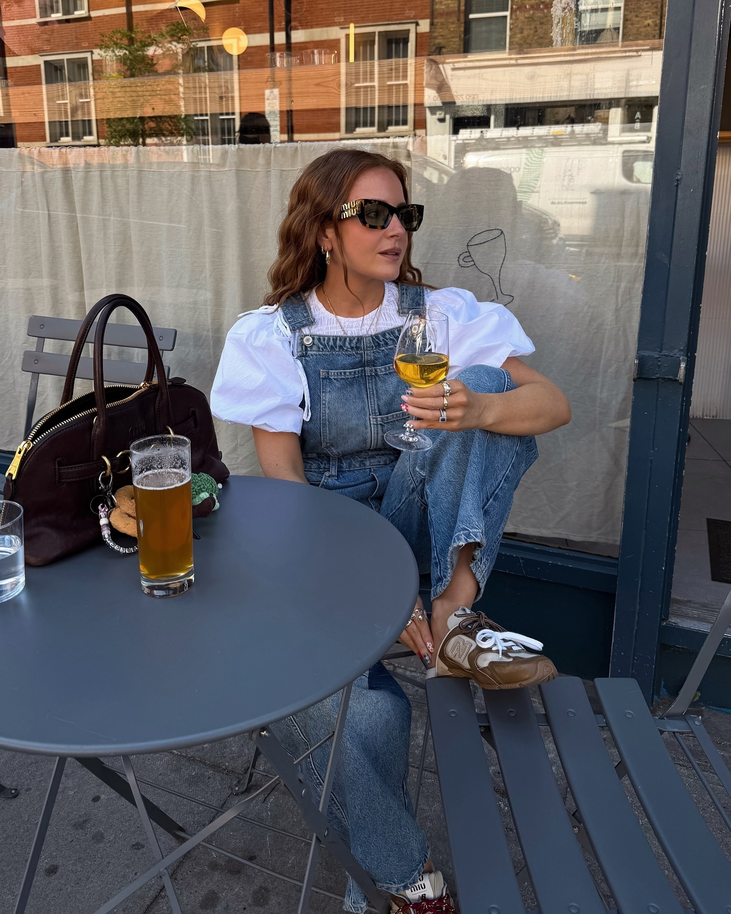 Influencer @rebeccaferrazwyatt sits at a table outside wearing Miu Miu X New Balance backless trainers with dungarees and a puff-sleeve blouse.