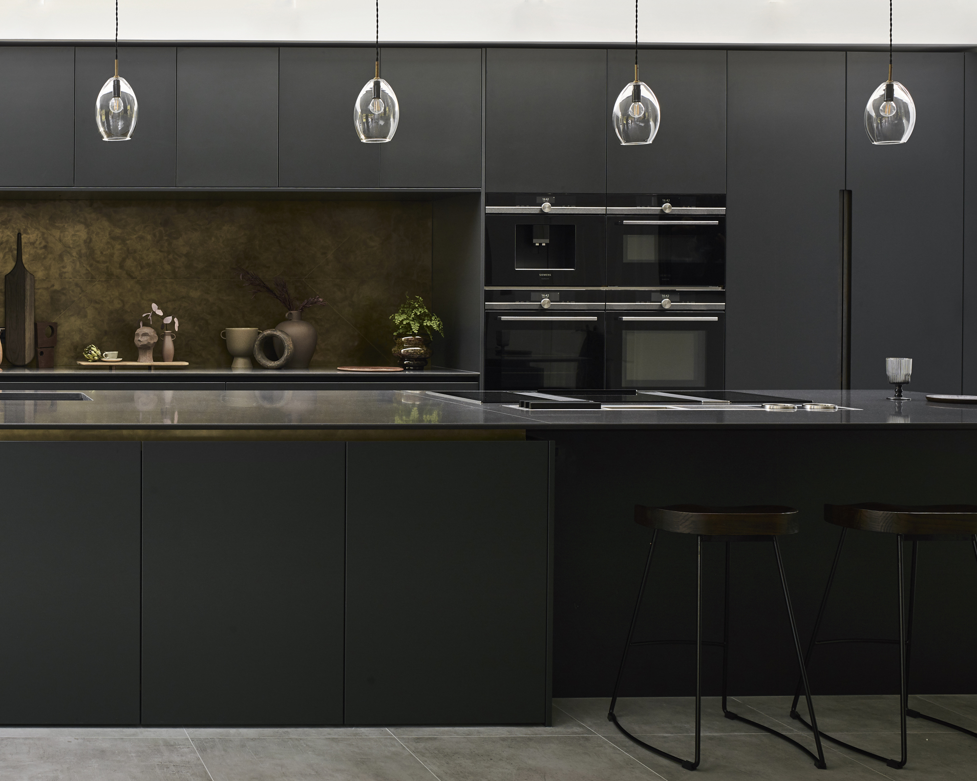 A black kitchen idea with matte black cabinets, stone worktop on the island, brass backsplash and pendant lights