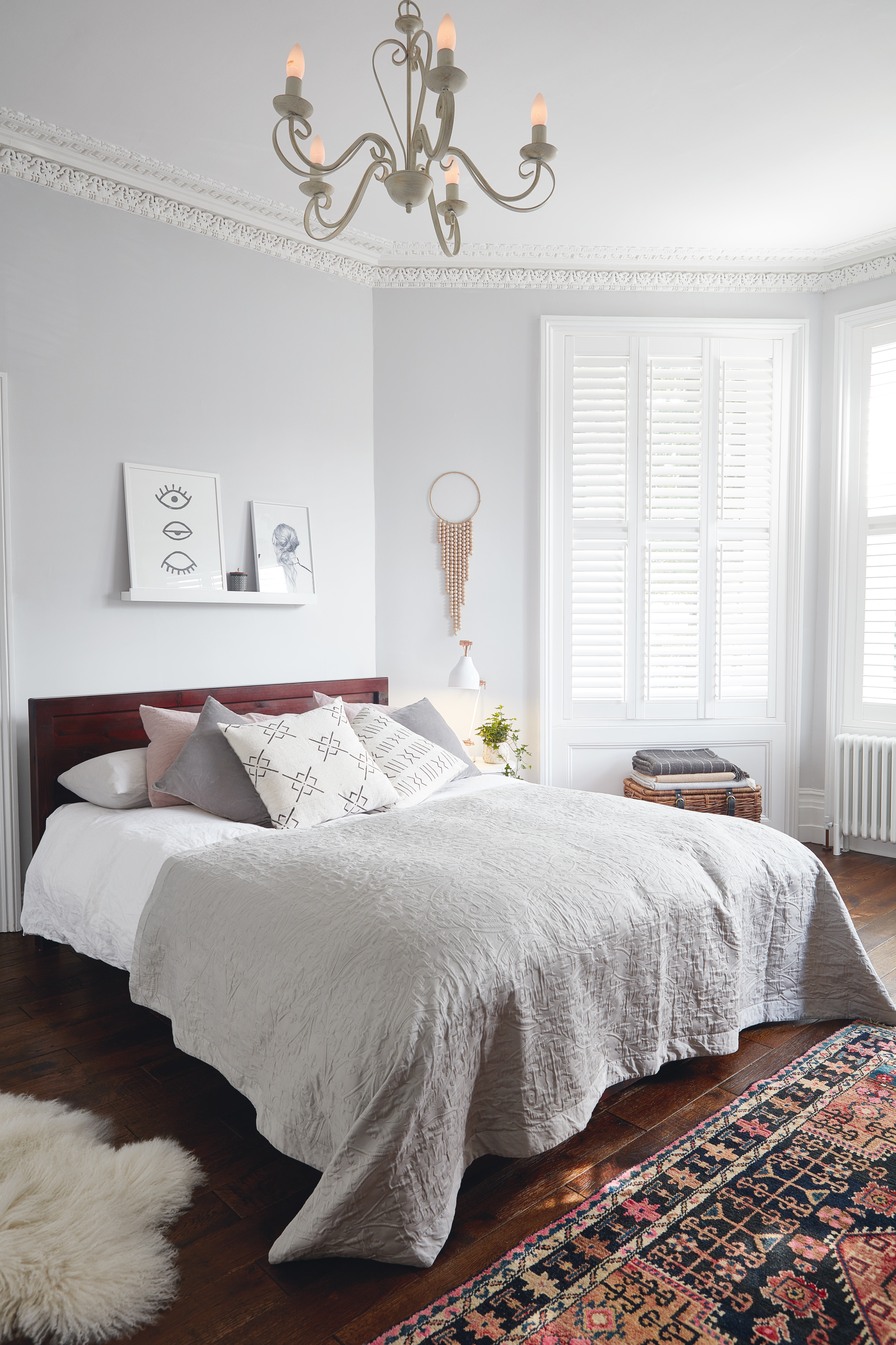 A bedroom with light grey wall paint decor, statement chandelier, shutter window treatment, floating shelves and ethnic rug