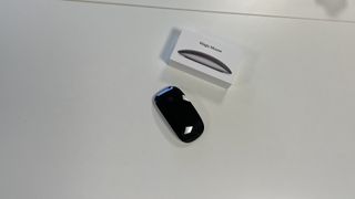 The black Magic Mouse on a desktop, placed near its box.