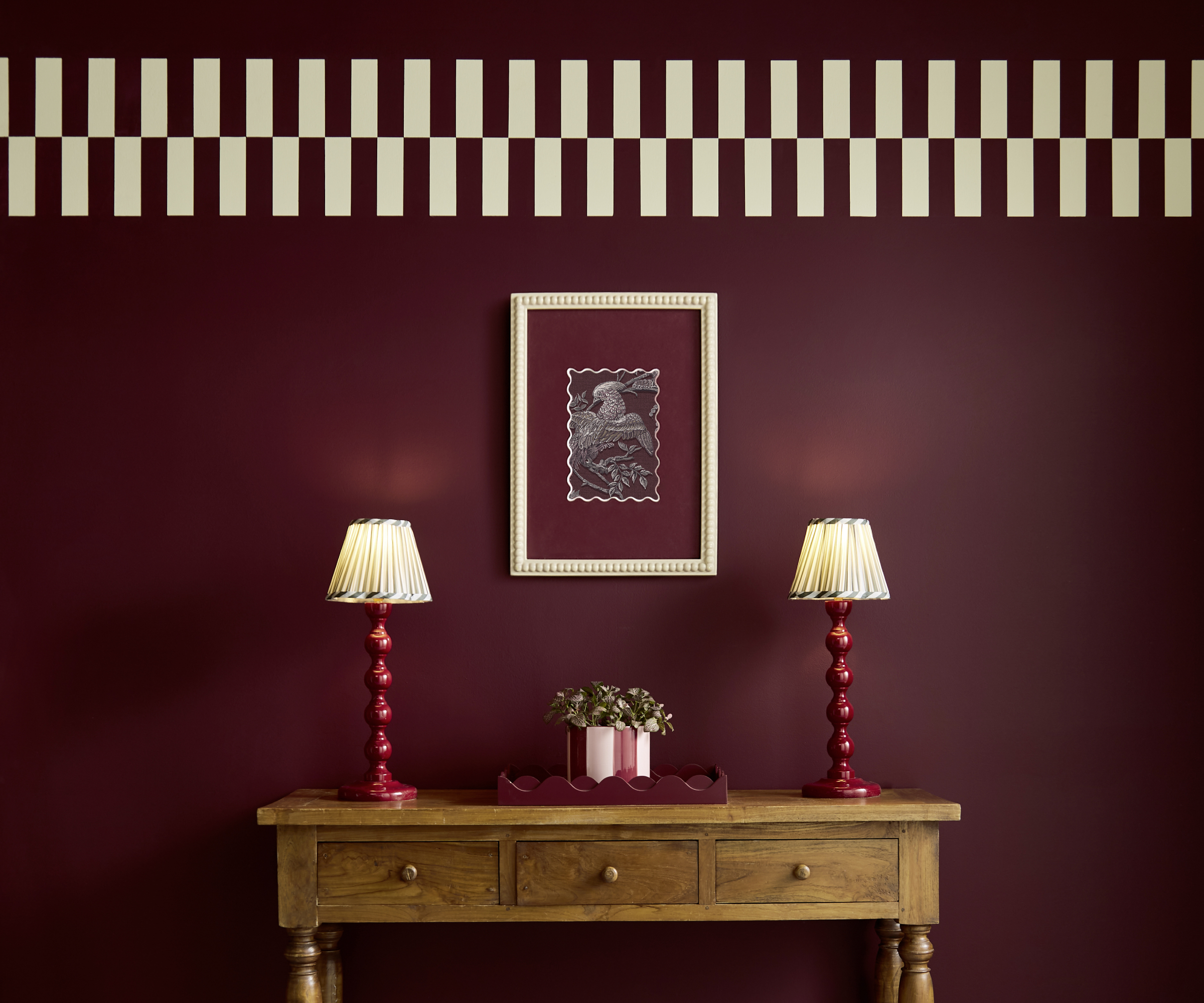 room with dark burgundy walls and cream and burgundy chequerboard border at top of wall, wooden console with table lamps