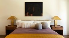 An off-white bedroom with a framed painting, a set of wood bedside tables, matching gold lamps, a trio of white pillows, two round gray pillows, and a purple and mustard bedscape