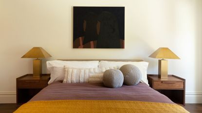 An off-white bedroom with a framed painting, a set of wood bedside tables, matching gold lamps, a trio of white pillows, two round gray pillows, and a purple and mustard bedscape