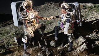 two people fist bump while wearing spacesuits.