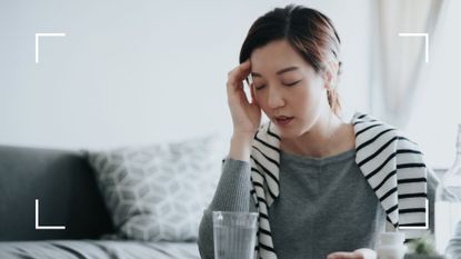 Woman experiencing keto headache with hand to head, next to a glass of water in light gray living room