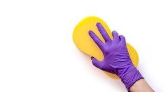 A surface being washed down with yellow sponge. The hand holding the sponge is wearing a purple kitchen glove.