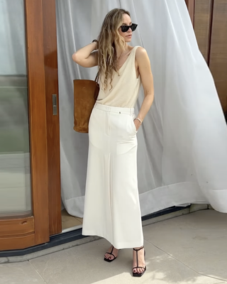 influencer Anouk Yve posing outside in a neutral tank top, white maxi skirt, and strappy sandals