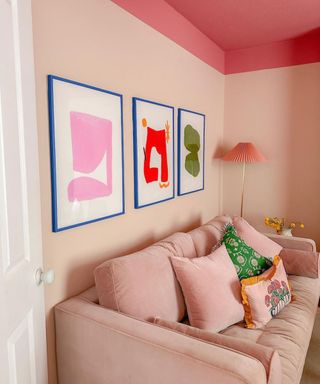 A pink living room with a pink couch with throw pillows on and a wall with three framed colorful wall art prints