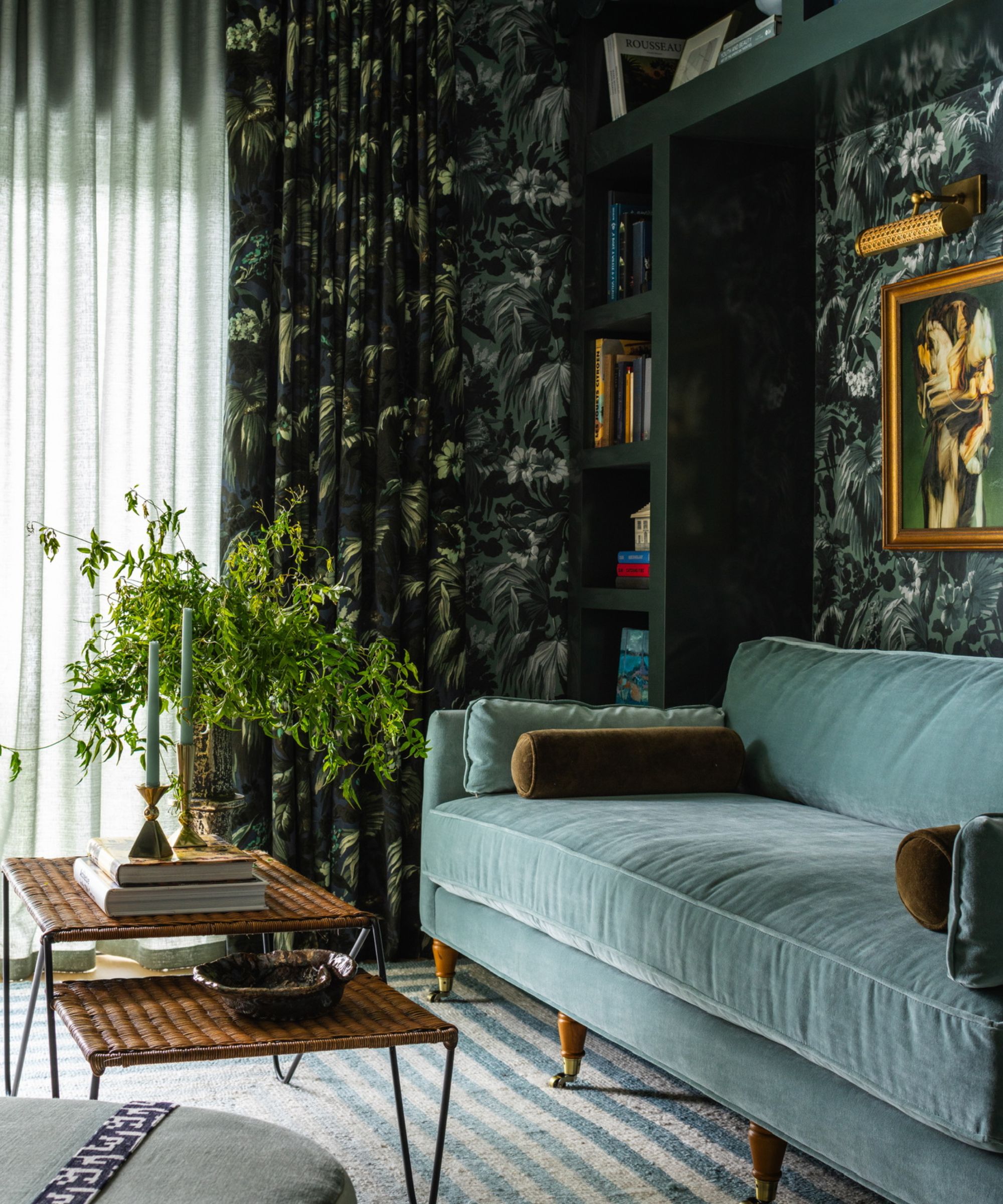 a sitting room with a teal couch, wallpapered walls, long drapery covering the windows, a rattan coffee table, and a plant against the wall