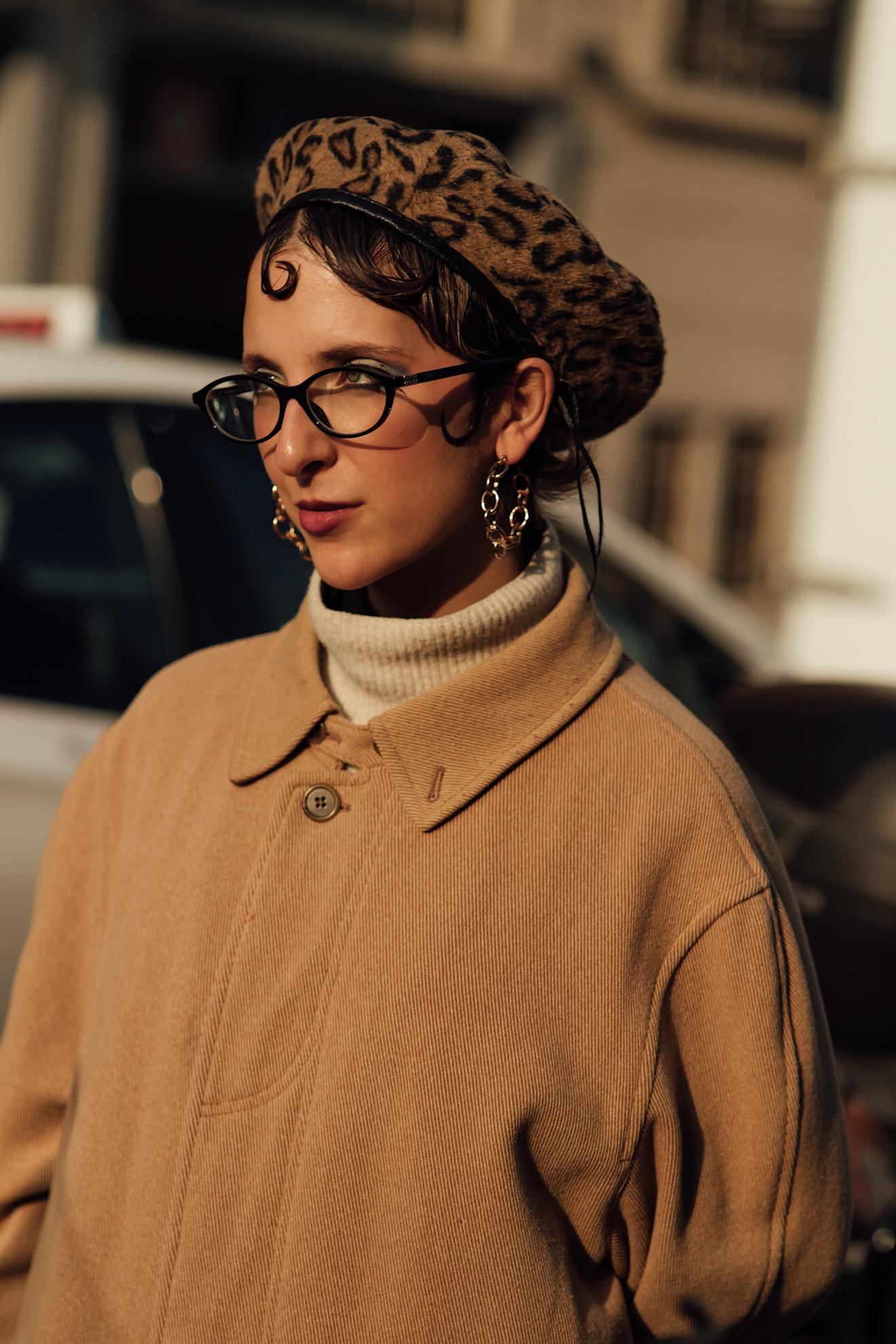 a woman in a leopard beret and tan jacket with a kiss curl hairstyle and glasses