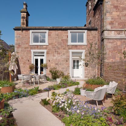 a house with garden in the foreground with a winding pathway, two seating areas and lots of flowers in purple and white