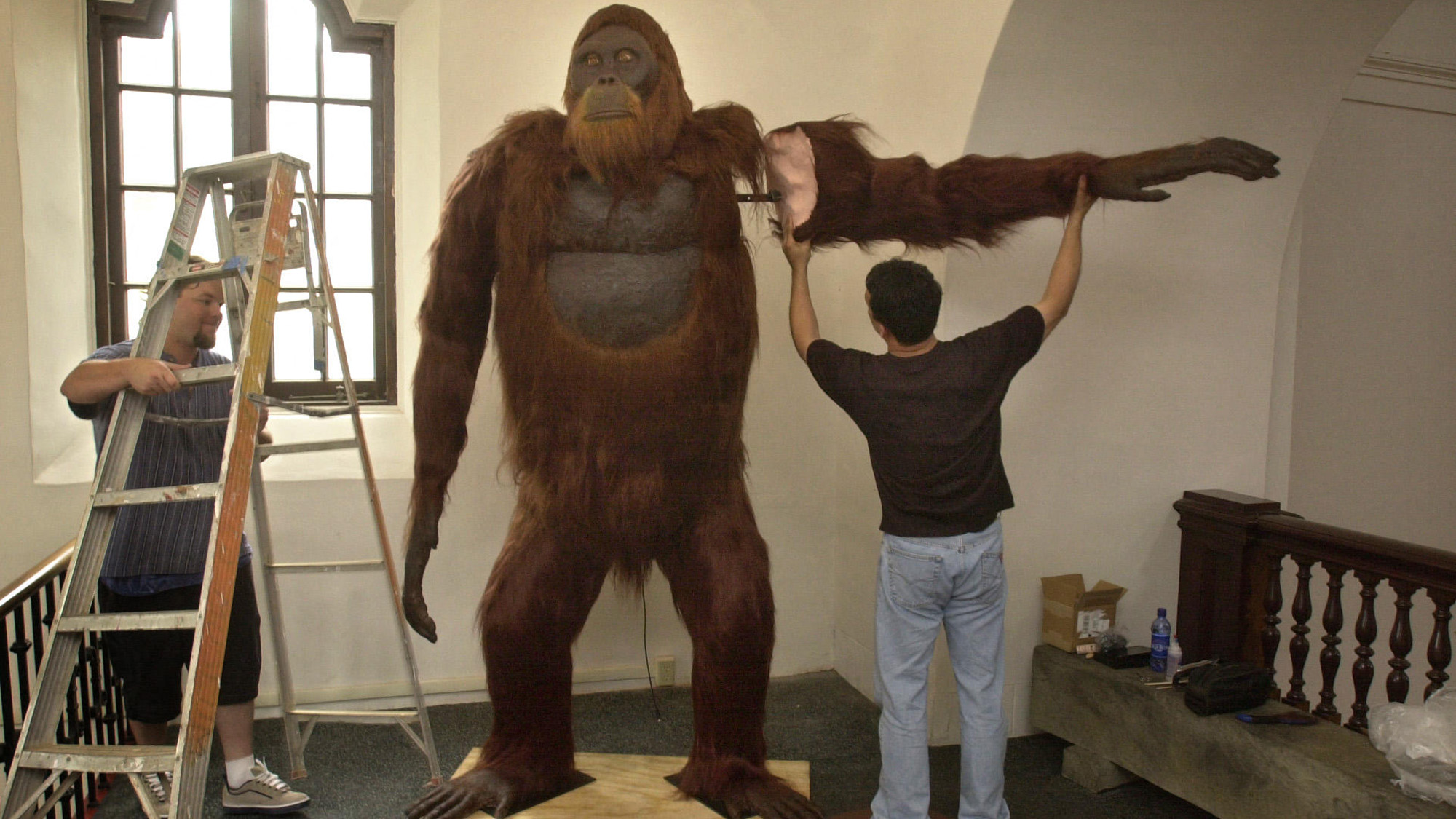 Two men stand next to a tall orange-furred ape replica in a museum.